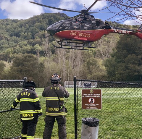 Two firefighters in uniform seen from behind looking toward a Contra Costa County Fire Rescue helicopter that is above a field