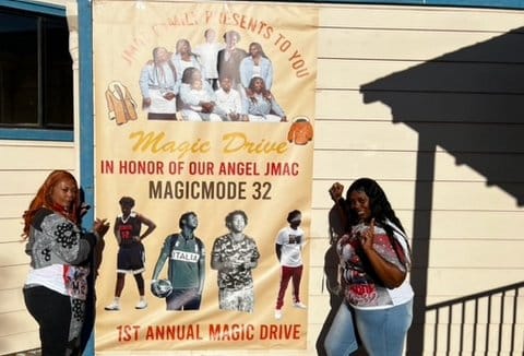 Two Black women standing next to a large poster with pictures of a Black teen boy and the words J Mac's family presents to you magic drive in honor of our angel J mac magic mode 32 1st annual magic drive