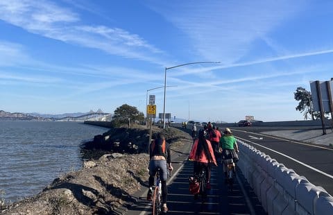 People biking on a roadway under an expansive blue sky with the San Francisco Bay to their left