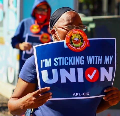 An older Black woman holding a sign that says "I'm sticking with my union." A man is out of focus behind her.