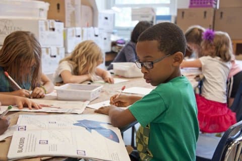 Young children sitting at a long table in a classroom. At the forefront is a Black boy with glasses.