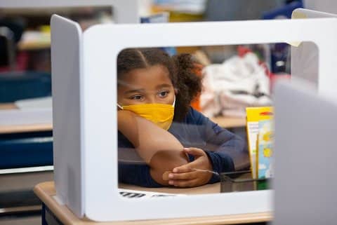 A young Black girl wearing a yellow mask sitting at her classroom desk, which holds a barrier with a clear front and white sides for COVID prevention. The girl looks upset or tired.