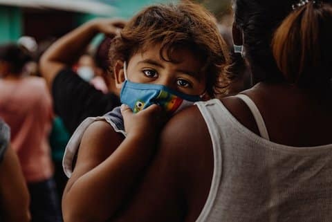 A small child being held looks over person's shoulder toward the camera. The kid is wearing a mask.