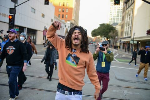 A Black man walks down a street amid other protesters with fist raised and mouth open.