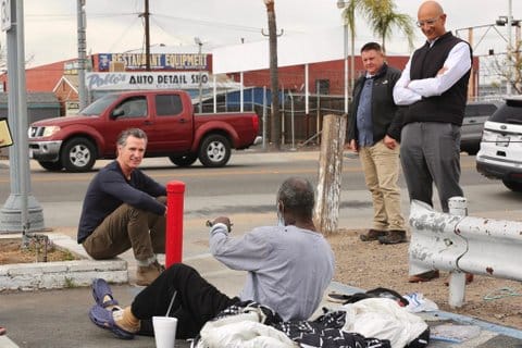 Gov. Gavin Newsom and an older Black man sitting on the ground near a street with two other men standing nearby
