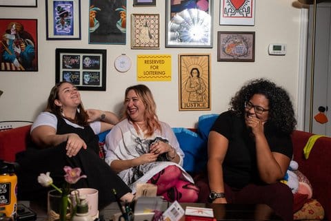 Two white women and a Black woman sitting on a couch, laughing or smiling. There are various artworks on the wall behind them.