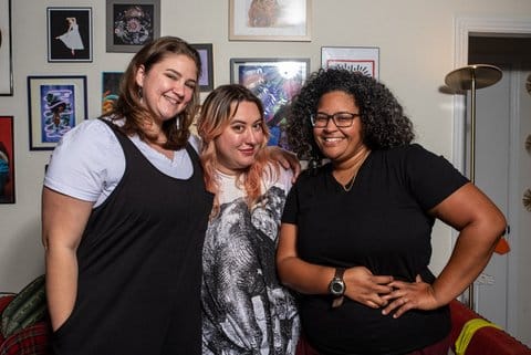 Two white women and a Black woman smiling standing shoulder to shoulder in their home