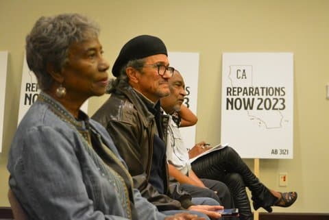 Side view of a row of people sitting. An older Black woman and two older Black men and a sign that says CA Reparations Now 2023 AB 3121 are visible.