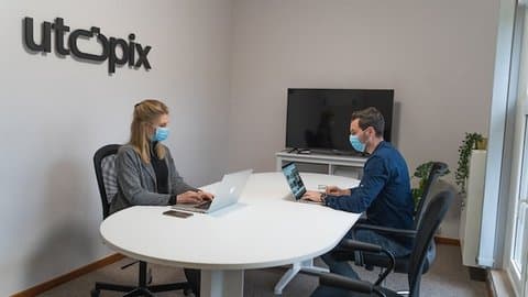 A woman and man sitting at a table, both wearing surgical masks and working on laptops. A large monitor is facing them, turned off, and a sign on the wall says utopix.