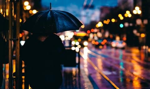 Person walking at night with an umbrella along a city street. Cars, lights and a bus are out of focus in the background.