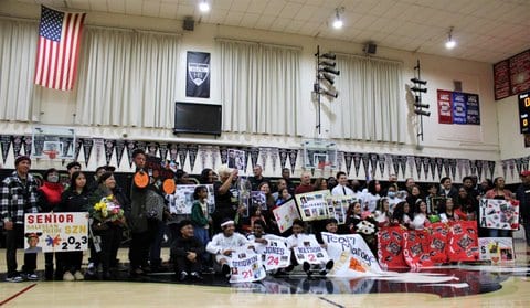 Crowd of adults and young people sitting or standing on the basketball court in a high school gym. Several are holding signs, including one that says Senior Salesian Pride Szn 2023.