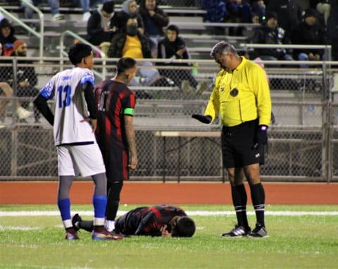One soccer player on the ground with two other players and a referee standing by