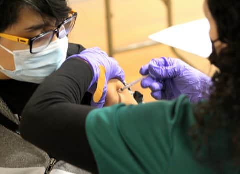 A nurse gives a teen boy a shot in his upper arm. The photo is taken from over the nurse's shoulder. Both are wearing masks, and the boy is looking at his arm as the shot is given.