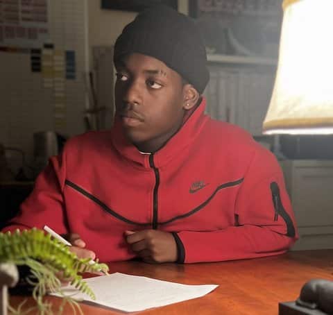 A Black teen sitting at a table with pen and paper. He is wearing a red Nike jacket and black beanie and looking to the side with a serious expression.