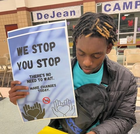 A Black teen boy holding up a poster that says "We stop you stop. There's no need to wait. Make changes today. The phrase "n-word" is written in a red circle with a line through it. There are drawings of black and white hands each holding a heart.
