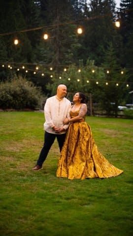 A man and woman standing together in a grassy area with lights strung above. They are looking at each other adoringly and both dressed nicely. She is wearing a marigold yellow dress with full skirt.