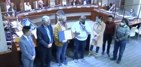 Screenshot showing seven people standing in a row at a government meeting. One person is holding an official proclamation. One has a microphone. One is Richmond Mayor Eduardo Martinez. Three are wearing masks.