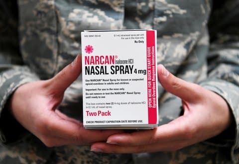 Box that says Narcan nasal spray. Use Narcan nasal spray for known or suspected opioid overdose in adults and children. The box is held by a person in a camouflage military uniform whose hands and chest are visible. 