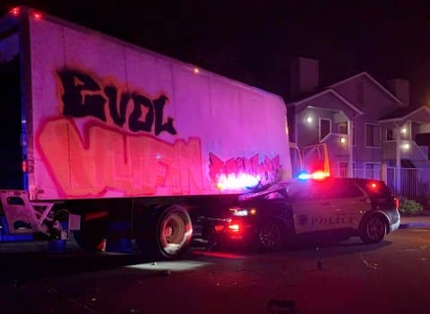 A white box truck at night that has crashed with a police SUV. The truck has graffiti on its side. The front of the cruiser is under the truck, and its siren lights are on, casting a colorful glow on the truck. A home is visible in the background.
