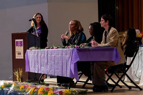 Three women sitting at a long table as speakers on a panel with another woman standing beside them at a lectern.