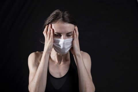 A white woman wearing a medical face mask has a distressed expression and is holding her hands to the sides of her head