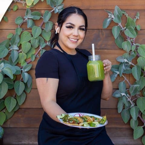 A smiling young Latina woman wearing a black apron and T-shirt holding a plate of tacos and a green beverage in front of a wood-paneled wall with leaves running down it.