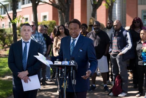 A Black man in a suit at a stand with TV news microphones with a white man in a suit next to him and a crowd of people behind him