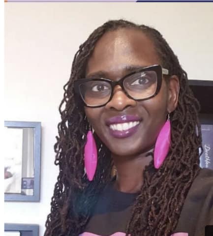 A smiling Black woman with long hair, black-framed glasses, purple lipstick and pink, feather-shaped earrings.