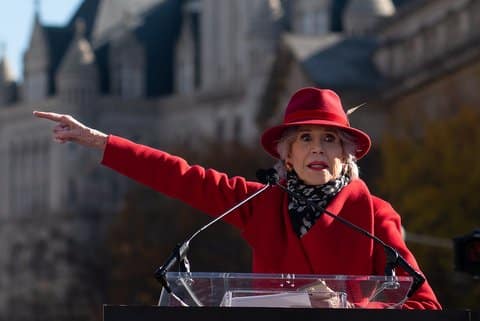 Jane Fonda wearing a red coat and hat standing at a lectern with microphones pointing left