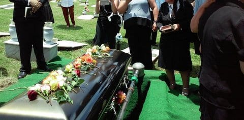 People standing around a casket adorned with flowers that has not yet been lowered into the grave. The photo is framed so that the people's heads are not visible.