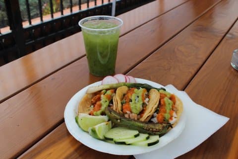 A plate of three tacos topped with tomato- and avocado-based salsas along with lime wedges, sliced radishes and cucumbers and grilled green onion and chili pepper. Alongside is a green iced beverage.