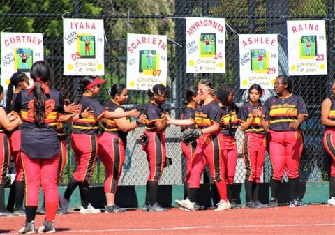Softball players lined up as teammate passes by to shake their hands. On a chain-link fence behind them are signs honoring senior players with the names Cortney, Iyana, Scarleth, Myrionnia, Ashlee, Raina.