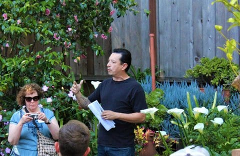 A Latino man speaks in front of a garden and wooden fence