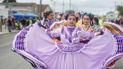 A row of Mexican dancers in colorful dresses led by a young woman fanning out her purple dress