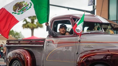 A man on the passenger side of an old school dark red Chevy 3100 pickup truck waves a Mexican flag as a young girl peeks over the car door