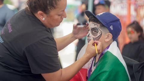 A boy wearing a Dodgers cap and a Mexican flag draped around his shoulders gets his face painted with a skull design by a woman