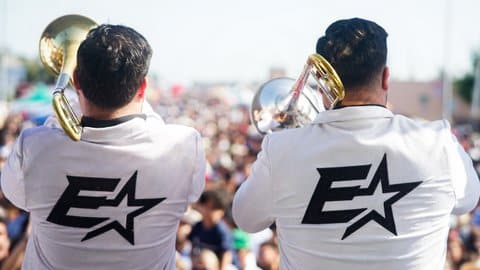 View from behind of two Hispanic men playing brass instruments and wearing white jackets with a black logo that features the letter E and a star