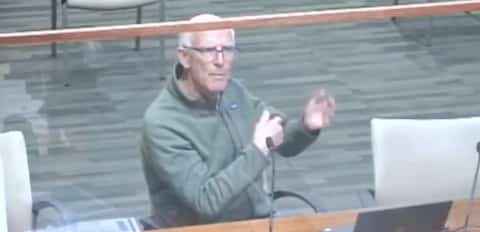 A balding white man with white hair gesturing with his hands as he speaks behind a clear barrier at a public government meeting