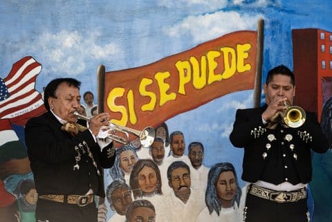 Two mariachi performers playing horns in front of a mural painted with brown-skinned people, a U.S. flag and a banner that reads Si, Se Puede.