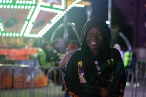 A smiling young Black girl in a jacket with the hood up in front of a brightly lit carnival ride
