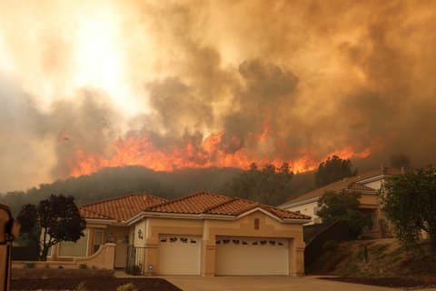 A house with a large wildfire burning in the hills behind it. The sky is smoky, yellowish, gray and dirty-looking.