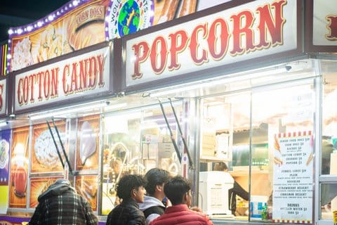 Three boys stand at a concession stand ordering window below large signs that say "cotton candy" and "popcorn"