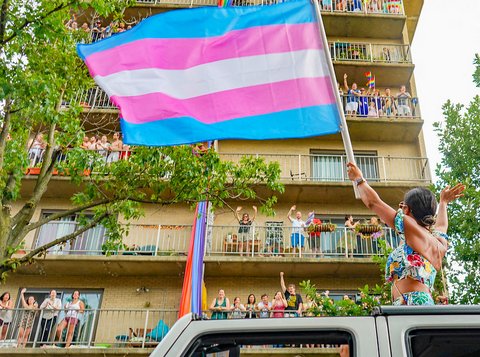 Looking up towards person waving transgender pride flag while standing in a car with the sunroof open. The person is facing a building with several people out on the balconies. Some are waving rainbow flags.