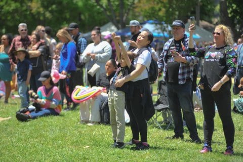 Among a crowd at a park event, a little boy stands with his arms in the air in front of a woman who has her arms around him