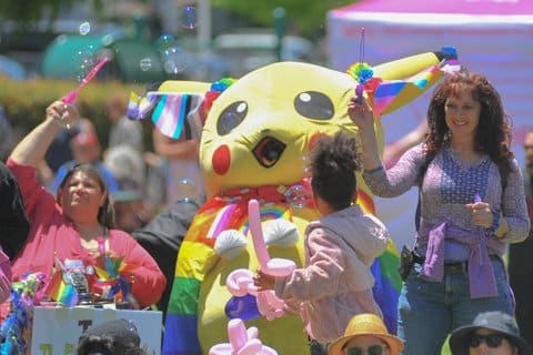 A person in a Pikachu costume with rainbow cape, a child with a balloon animal and two women with bubble wands