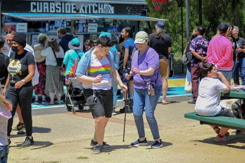 A person in a white T-shirt with a rainbow pattern across the chest and another person dance. Behind them is a food truck called Curbside Kitchen