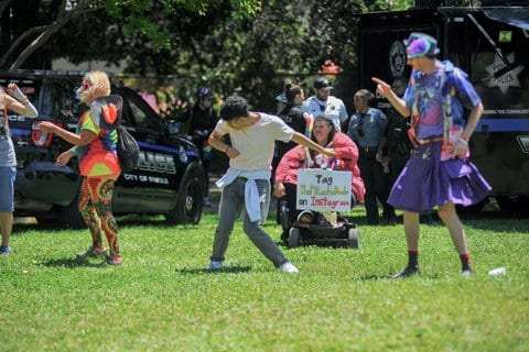 A young man dances on grass. Around him are colorfully dressed people and a person on a motorized wheelchair with a sign that says tag ThePikachuDude on Instagram