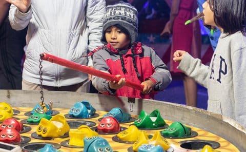 A small Hispanic boy playing a carnival game holds a rod with a chain and hook at the end over plastic frogs, some with their mouths open