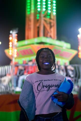A young Black girl stands in front of a colorful tower drop ride. She is wearing something glow-in-the-dark on her teeth and a pink shirt that says "I'm not perfect but I'm original" and holding a stuffed animal.