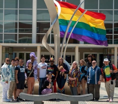 Fifteen people, one of whom is holding a small dog, in front of a rainbow flag